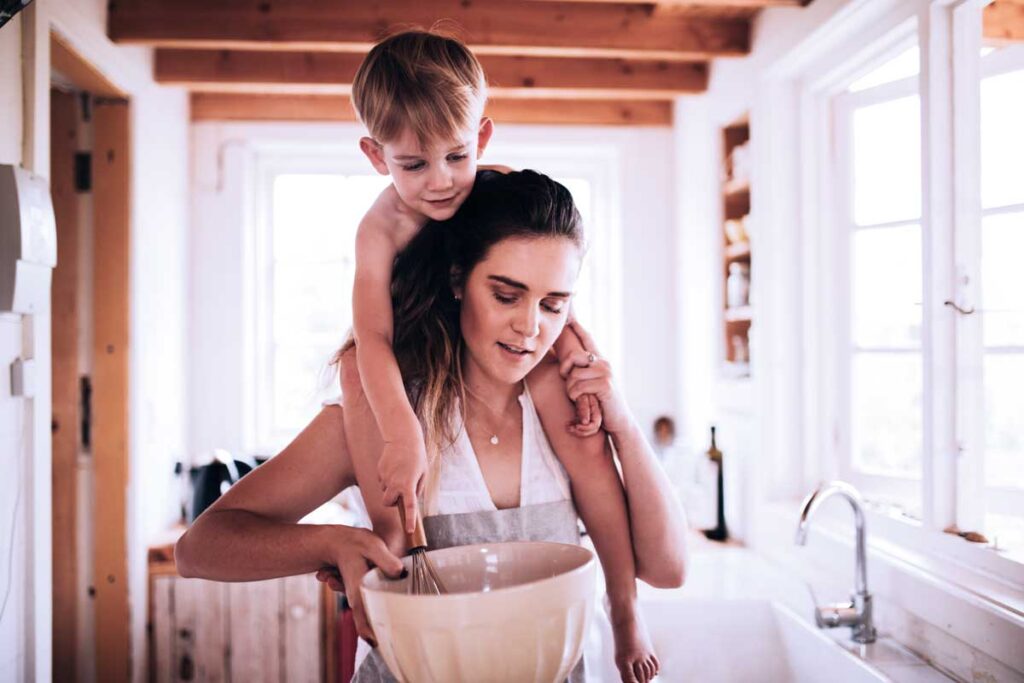 Busy mom in a farmhouse kitchen with her son on her shoulders. They are stirring a glass bowl with a whisk