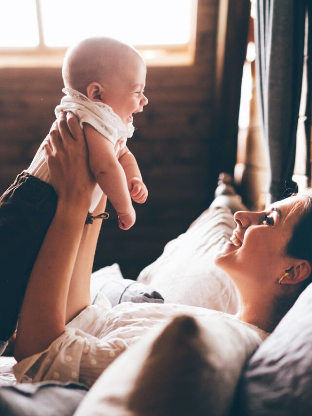 mom holding her baby up over her, smiling while laying in bed