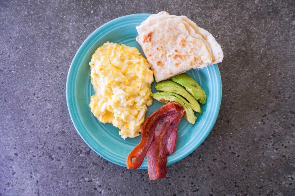 top view of a breakfast plate with cottage cheese eggs, sourdough tortilla, bacon and avocado on a concrete countertop