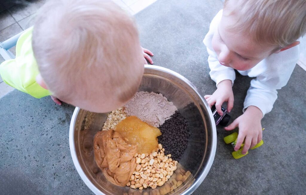 two blonde boys looking into a stainless steel mixing bowl with peanut butter, peanuts, chocolate chips, honey and protein powder