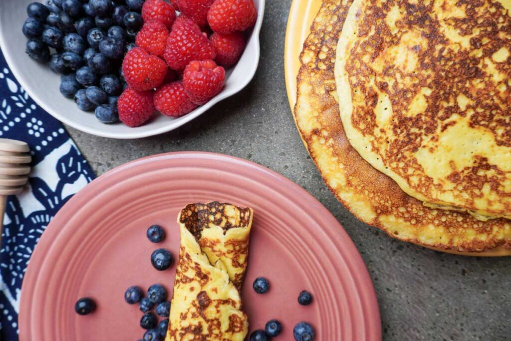 One rolled up crepe next to a plate of crepes and fresh raspberries and blueberries