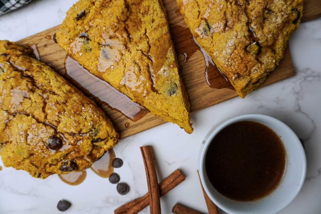 Close up of chocolate chip pumpkin scones on a wooden board with a bowl of maple vanilla glaze