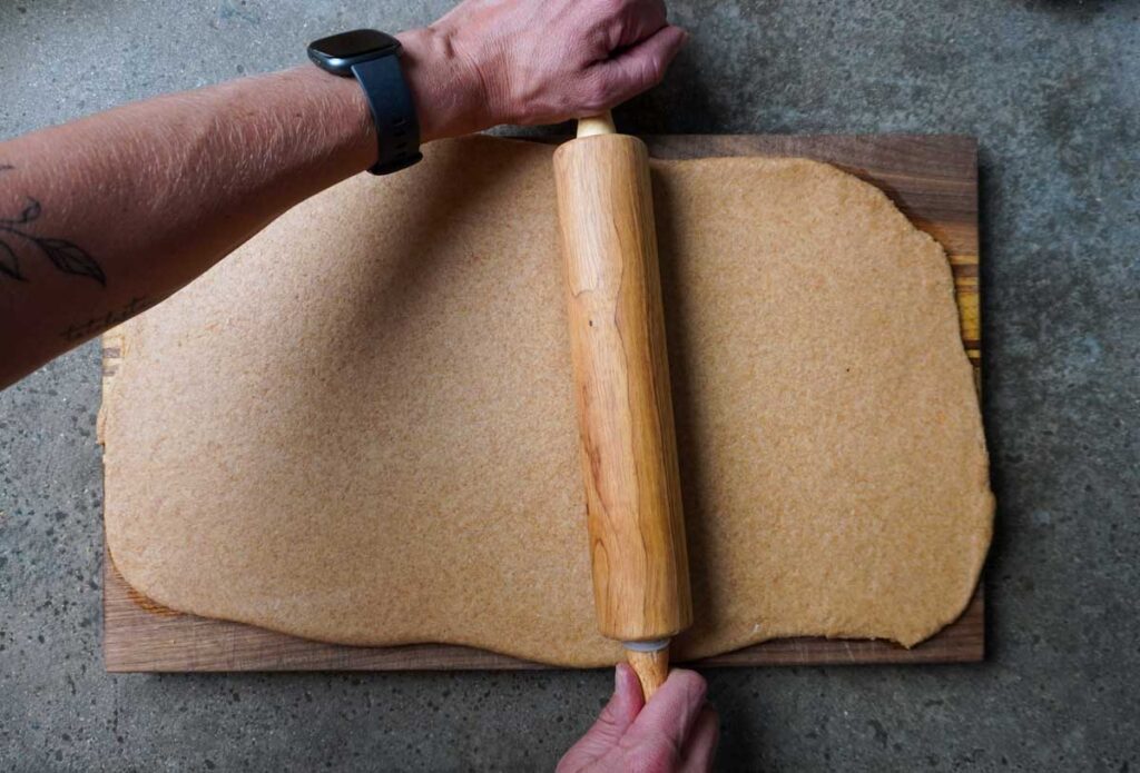 Top view of two hands rolling out fresh milled flour dough onto a cutting board.